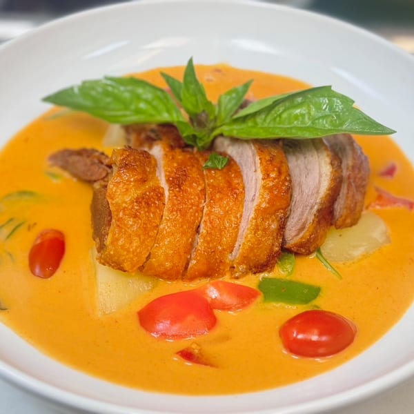 Sliced crispy duck in red curry with cherry tomatoes, pineapple, and basil in a white bowl.